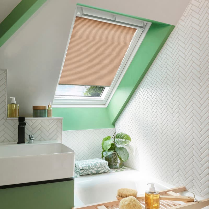 Beige pleated skylight blind, lowered to cover most of a roof window and filtering daylight; in a sloped, greenâ€‘accented bathroom above a bathtub with white herringbone tiles, plant, sink.