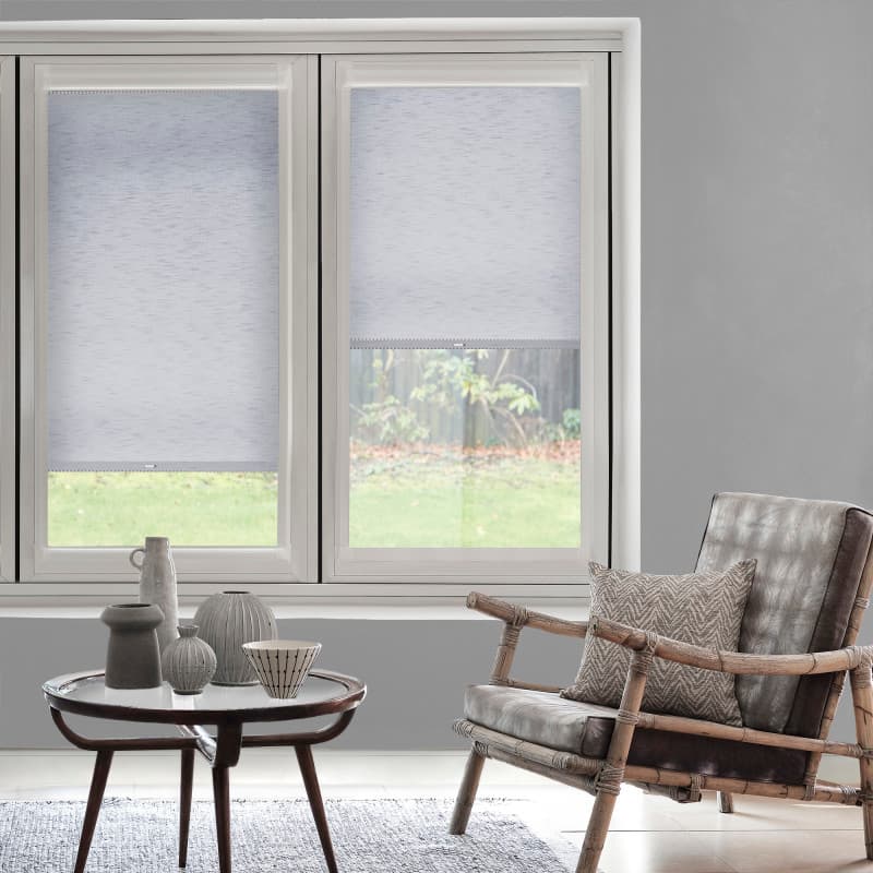 Light-gray roller blinds cover twin tall windows â€” left fully lowered, right half-raised â€” softly filtering daylight over a living room with a wooden armchair, small table with ceramics, and a grassy garden view.