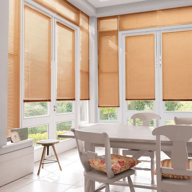 Beige pleated blinds covering tall bay windows and French doors, fully lowered and filtering warm daylight; bright dining nook with white wooden table and chairs, floral cushions, and garden view below.