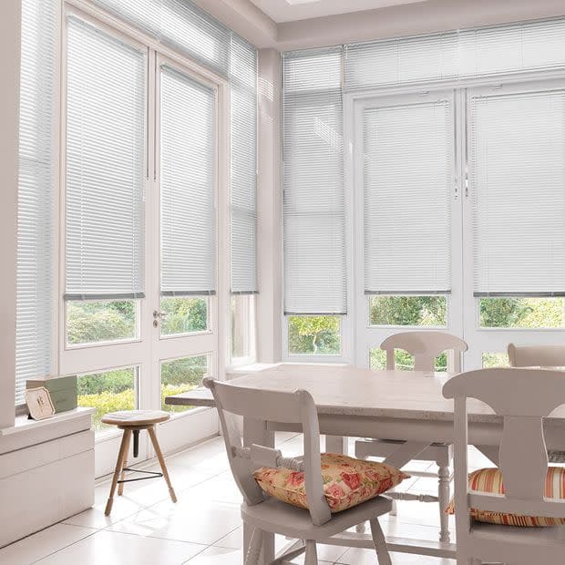White horizontal Venetian blinds lowered across tall bay windows, slats mostly closed and softly filtering daylight, in a bright dining sunroom with white table, chairs, cushions and garden view.