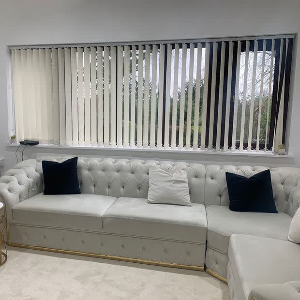 Cream vertical blinds across a wide window, darker slats at right, drawn and tilted to partly reveal a garden; living room with pale grey tufted sectional and dark cushions.
