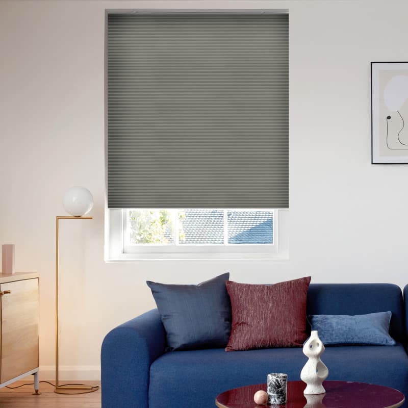 Gray pleated cellular blind lowered over a recessed window, blocking light with a narrow gap at bottom; modern living room context with blue sofa, colored cushions, floor lamp, and daylight.