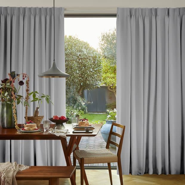 Object: light-gray, floor-to-ceiling pinch-pleat curtains parting at center; Action: framing a bright opening and admitting diffuse daylight; Context: wooden dining table set for breakfast, pendant lamp, garden view.