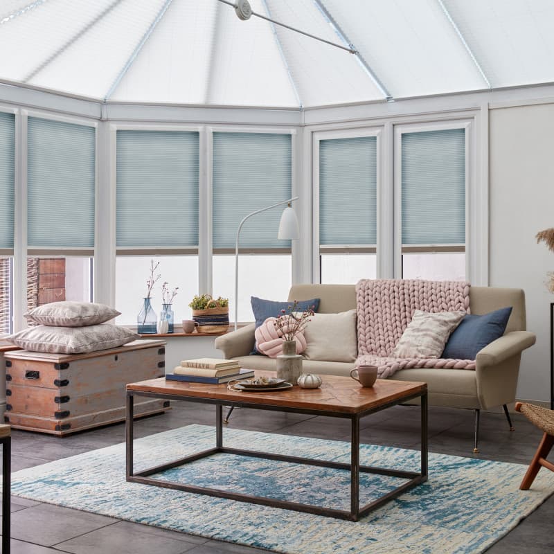 Pale blue cellular (pleated) blinds lowered across bay windows and conservatory roof, filtering soft daylight; in a sunlit living conservatory with beige sofa, pink throw, wooden coffee table, patterned rug.