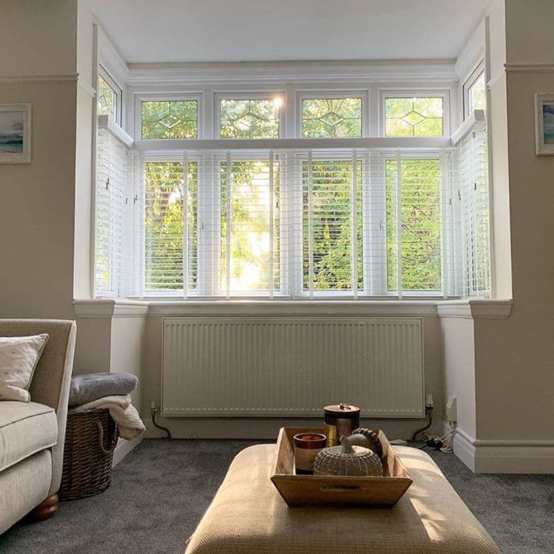 White slatted horizontal blinds covering a threeâ€‘pane bay window with decorative transom panels, partially open and filtering warm sunlight and leafy garden view into a neutral-toned living room with ottoman.