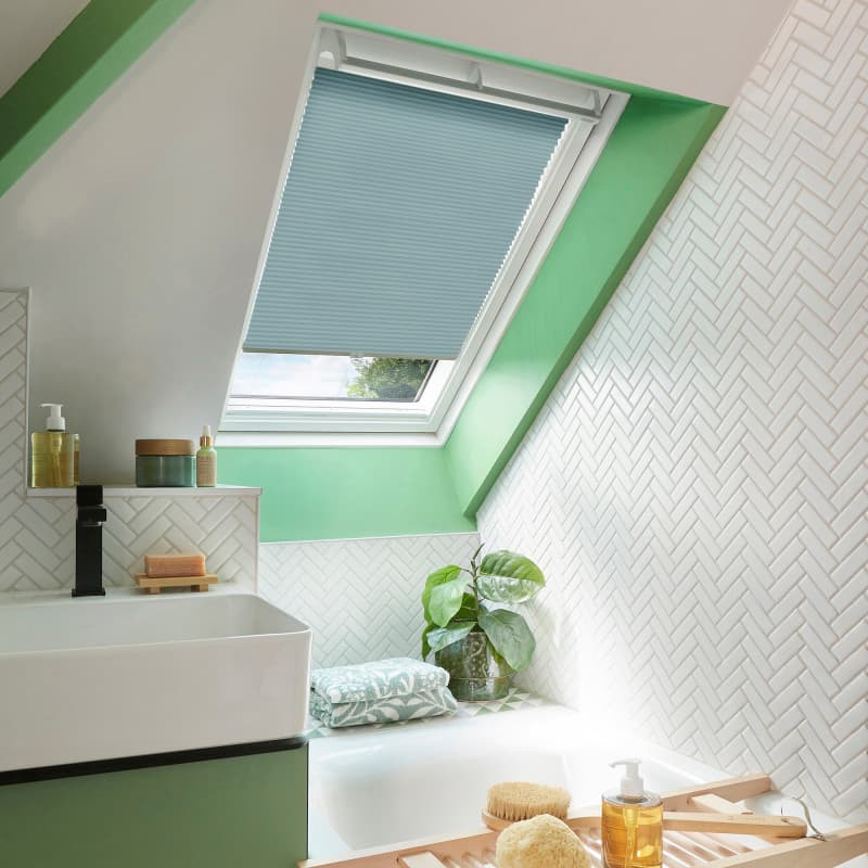 Light-blue pleated skylight blind, lowered halfway and filtering soft daylight; in a sloped attic bathroom with green-painted trim, white herringbone tiles, a basin, bathtub, and a potted plant.