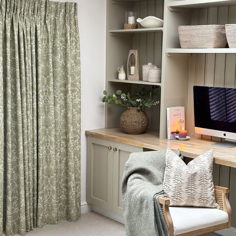 Pleated sage-green floral curtains, full-length and drawn closed, gently filtering daylight; beside a neutral built-in desk with wicker vase, iMac, lit candle, basket shelves, and a chair with throw.

Text: WHITE