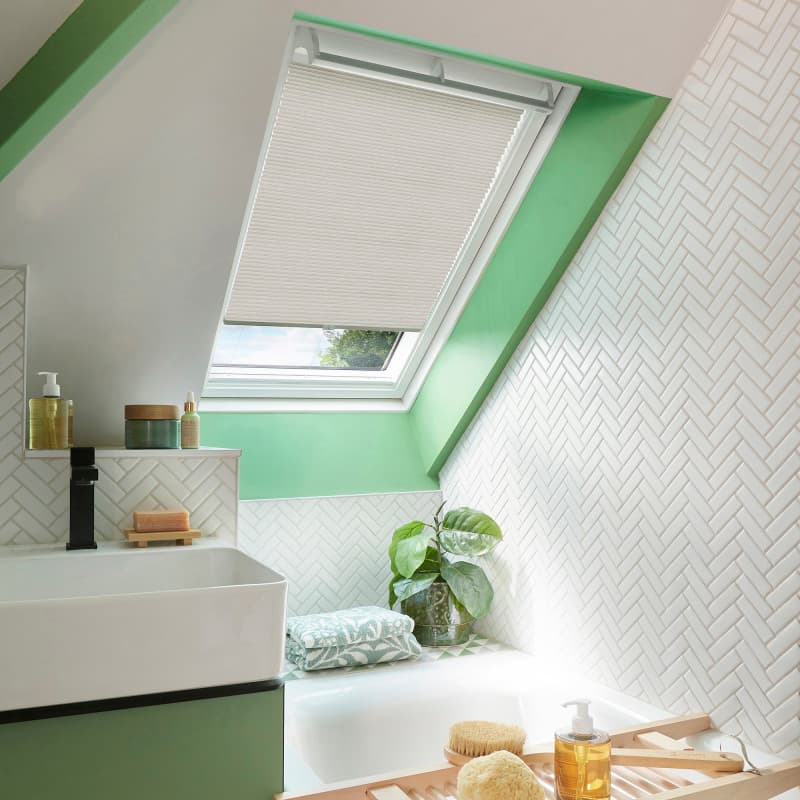 Cream pleated skylight blind, mounted on an angled roof window, partially lowered and filtering daylight; in a bright attic bathroom with green window trim, white herringbone tiles, sink, tub, plant.
