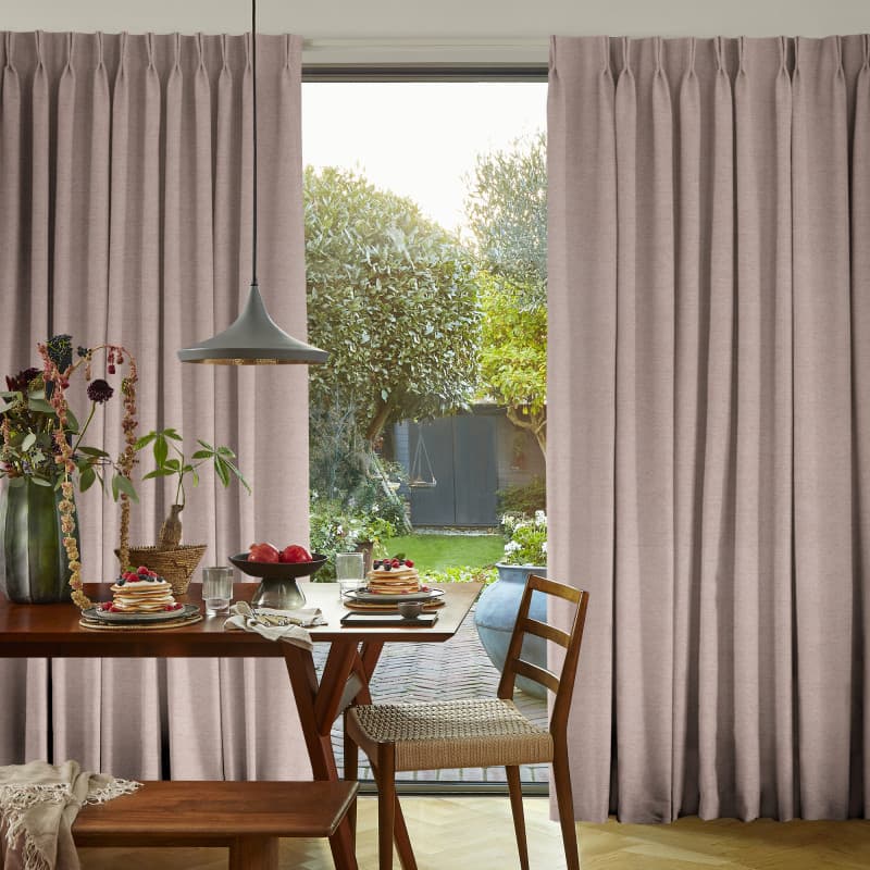 Beige pinch-pleat floor-to-ceiling curtains, parted centrally, framing and filtering soft daylight through a glass sliding door; a dining table set for breakfast and chairs in a sunlit, garden-facing room.