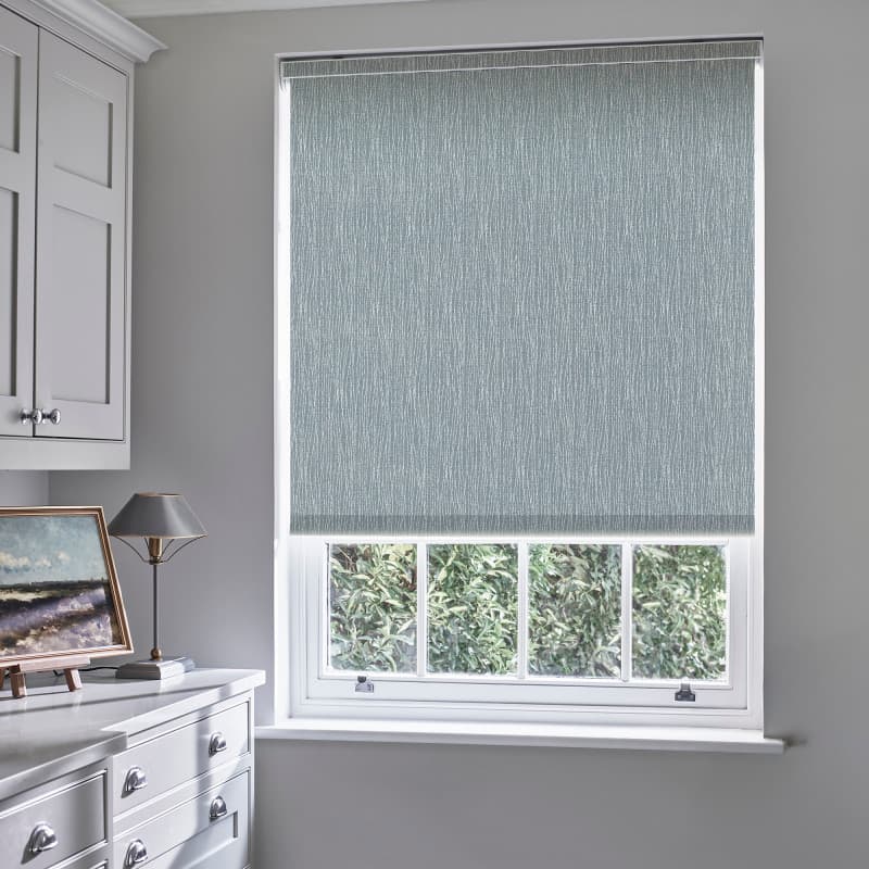 A blue-gray textured roller blind covers most of a sash window, lowered to filter light; surrounding neutral kitchen with white cabinetry, lamp, countertop and leafy exterior visible below.