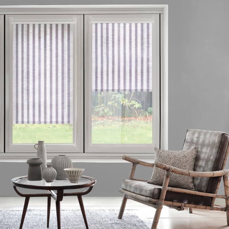 Striped fabric roller blinds (white with narrow vertical gray stripes), partially lowered, filtering soft daylight in a living room with a rattan armchair, coffee table and grassy yard visible outside.