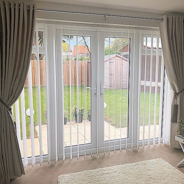 Beige curtains tied back frame white vertical blinds across patio doors, filtering daylight and revealing a small fenced backyard with lawn and a wooden garden shed.