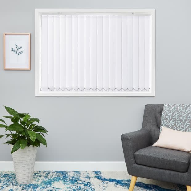 White vertical blinds filling a recessed window, closed and filtering daylight; set above a pale grey living room wall with a grey upholstered chair, cushion, potted plant and blue rug.