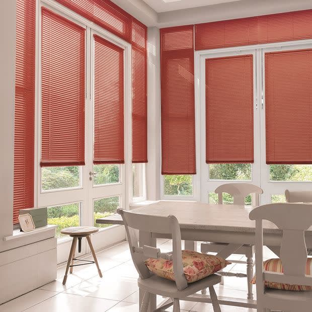 Red horizontal slatted blinds covering tall bay windows and doors, mostly lowered and filtering daylight; inside, a bright dining nook with white table, chairs, tiled floor and glimpses of garden.