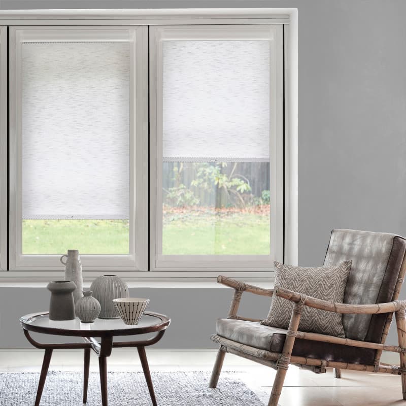 White textured roller blinds, left fully lowered and right partially lowered, filtering daylight over two tall windows; contemporary gray living room with bamboo-frame leather armchair, coffee table, and garden view.