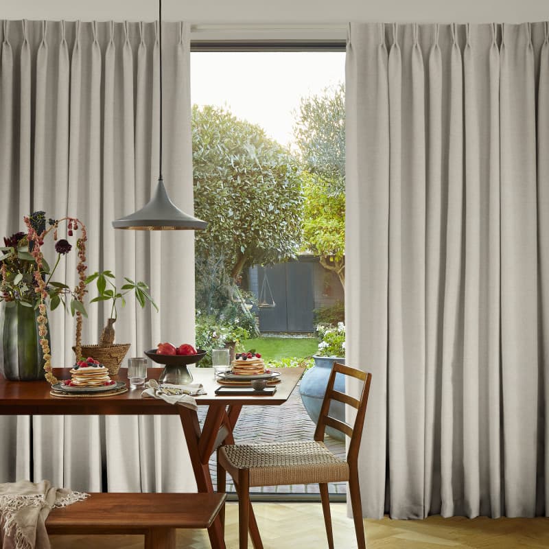 Beige pleated full-length curtains, centrally parted with soft folds, framing a glass patio door; they reveal a sunlit backyard while a wooden dining table set for breakfast sits inside.