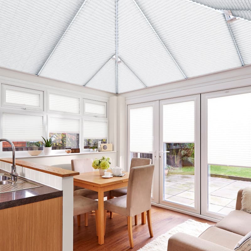 Object: white pleated blinds covering windows, French doors and pitched conservatory roof. Action: lowered, filtering and diffusing bright daylight. Context: airy kitchen-dining conservatory with wooden table and patio garden view.