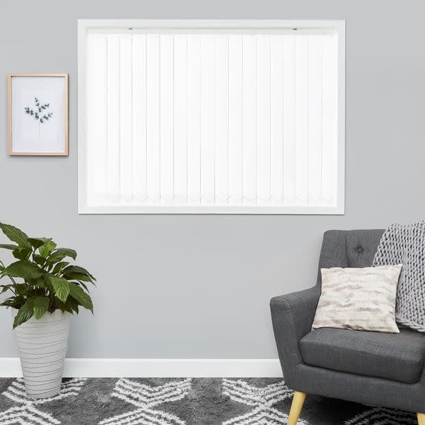 White vertical blinds covering a window, closed and diffusing daylight; in a gray living room with a potted plant, framed botanical print, gray armchair with cushion and patterned rug.