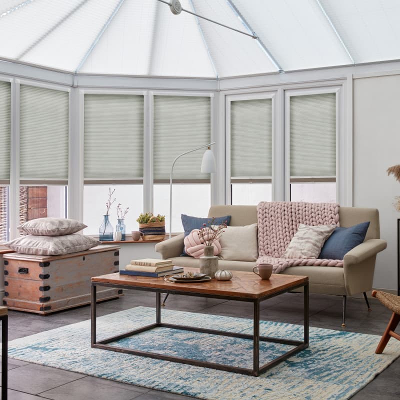 Pale green pleated cellular blinds cover tall conservatory windows, lowered mid-height and softly filtering daylight for privacy; framed by a beige sofa, wooden coffee table, and blue patterned rug.