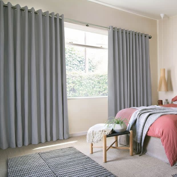 Grey grommet-top, floor-length curtains, pleated and hung on a metal rod, drawn open at center to frame and softly filter daylight through a window showing a leafy hedge; sunlit bedroom interior.