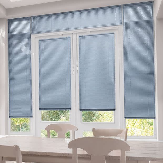 Light-blue horizontal venetian blinds span five tall window panels and a transom, lowered to cover upper portions while revealing narrow bottom panes; dining table and chairs, soft daylight, garden view.