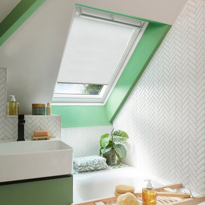 White pleated skylight blind, mostly lowered over a roof window, filtering daylight; bathroom with green-painted sloped ceiling, white herringbone tiles, a sink and tub, plant, and toiletries.