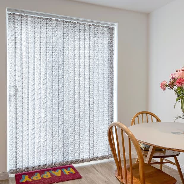 Patterned vertical blinds, full-height, drawn closed over a sliding door, softly filtering daylight; nearby round dining table with wooden chairs and pink flowers on laminate floor. Text: HAPPY