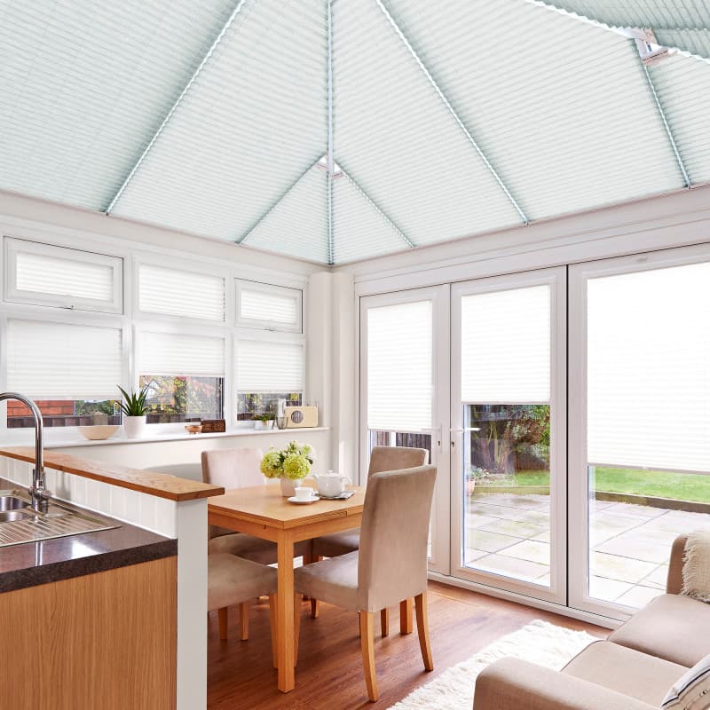 Pleated white conservatory blindsâ€”roof panels and window/door coveringsâ€”closed and filtering soft daylight; in a bright dining conservatory with wooden table, beige chairs, kitchen sink, and patio garden beyond.