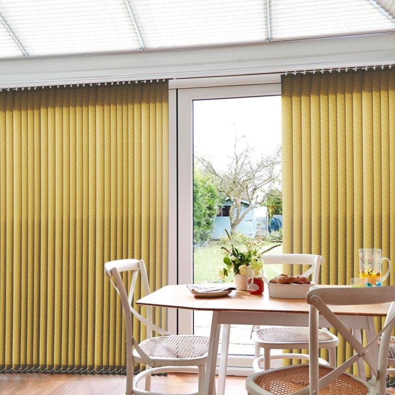 Yellow vertical blinds, mostly closed with a central gap, filtering daylight and revealing a backyard tree and shed through a glass door, in a dining nook with table and chairs.