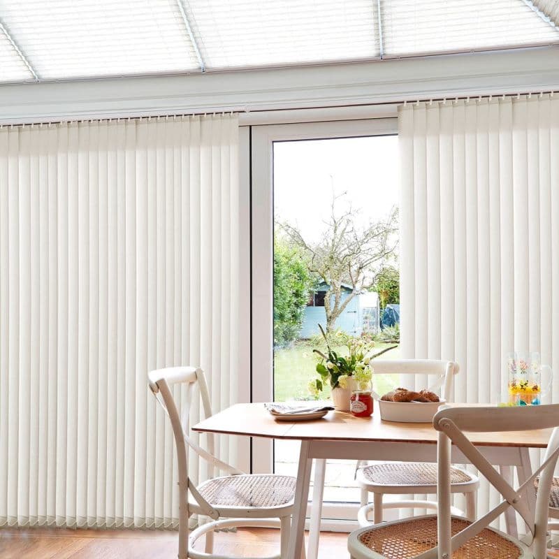 Cream vertical blinds span a large sliding glass door, mostly closed but parted centrally to reveal a sunlit garden; modern dining area in front with wooden table, wicker-seated chairs, flowers.