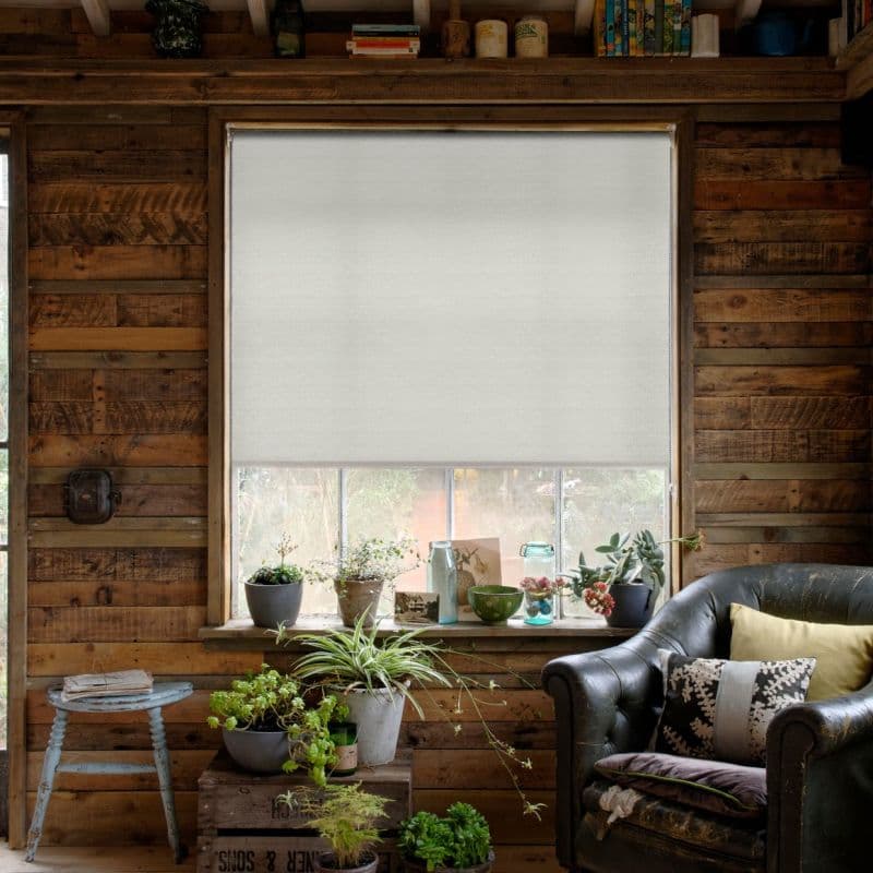 White fabric roller blind lowered inside a wooden frame, semi-opaque with a narrow bottom gap; filtering soft daylight into a rustic plank-walled room with potted plants on the sill and a leather armchair.