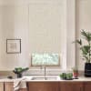 Patterned roller blind covers a kitchen window, filtering daylight; stainless faucet and double sink sit below on a marble countertop, flanked by potted plants, a bowl of greens, and framed sketch.