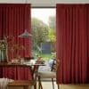 Deep red pleated floor-length curtains, split centrally and textured; partially drawn, framing and filtering daylight; dining room with a table set under a pendant lamp, opening to a green garden.