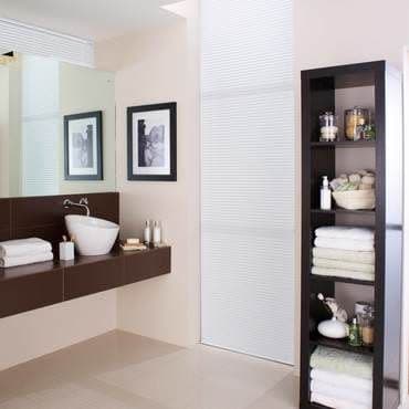 White pleated blind covers a tall narrow window, closed and diffusing daylight; modern beige bathroom with floating dark vanity, vessel sink, mirrored wall, framed art, and open shelving of towels.