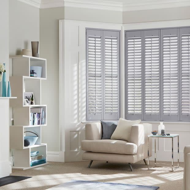 White plantation shutters on a bay window, closed with angled slats filtering sunlight into the room; neutral living room with beige armchair, throw pillows, open shelving and side table.