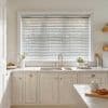 White horizontal slatted blinds across a large window, lowered and slightly tilted to filter soft daylight; set above a farmhouse-style sink in a bright, neutral-toned kitchen.