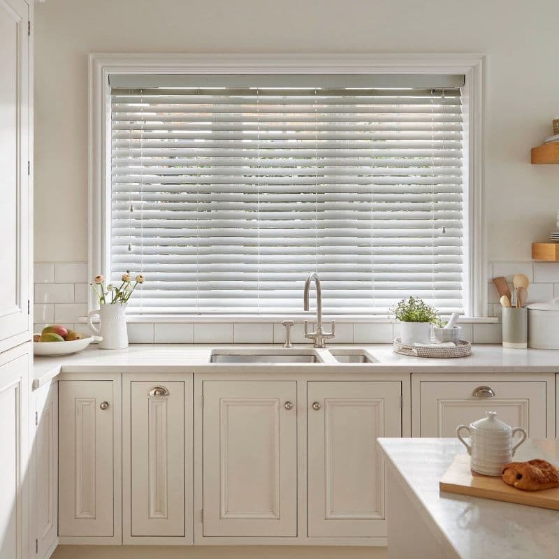 White horizontal slatted blinds across a large window, lowered and slightly tilted to filter soft daylight; set above a farmhouse-style sink in a bright, neutral-toned kitchen.