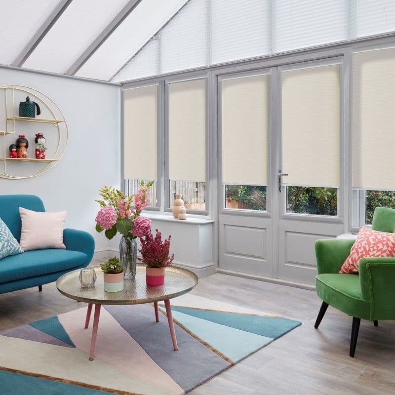 Beige roller blinds lowered across tall glazed French doors and windows, light‑filtering opaque fabric leaving narrow lower gaps; a bright conservatory-style room with colorful sofas, rug and flowers.