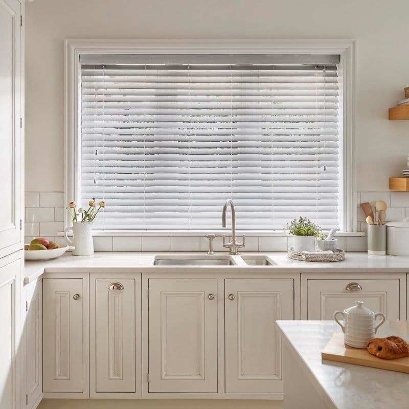 White horizontal Venetian blinds cover a large window, lowered with slats tilted to softly filter daylight; they sit above a farmhouse sink in a bright, neutral kitchen with cream cabinets.