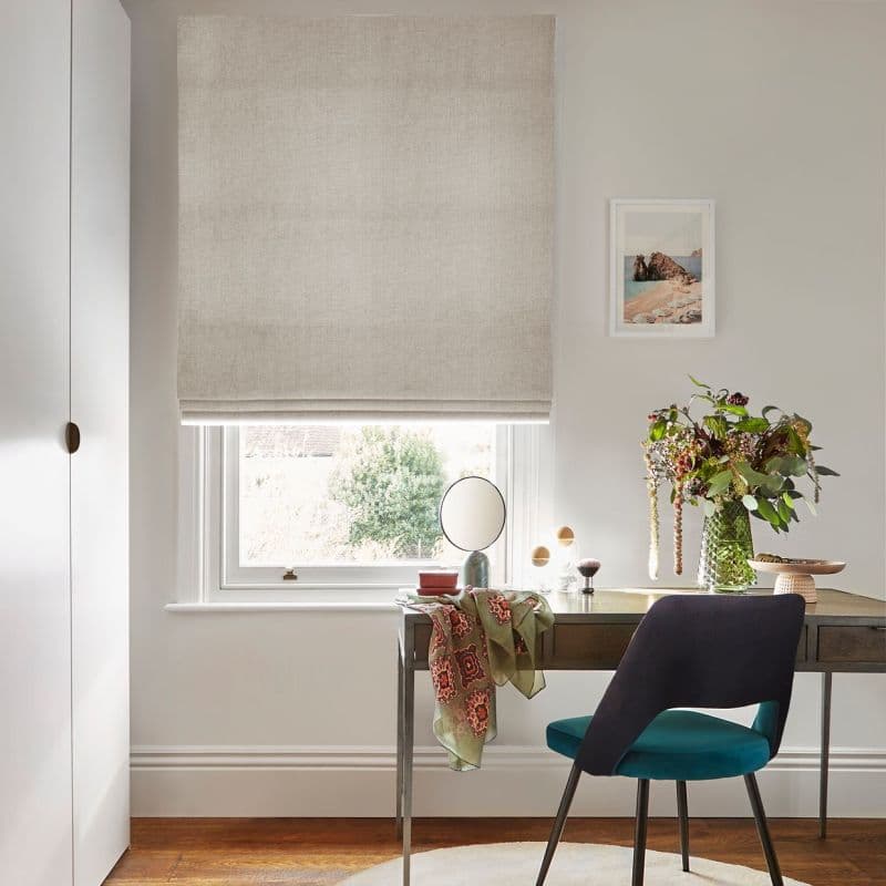 Beige linen Roman blind, lowered with a small fold, filtering soft daylight from a partially visible window; a desk below holds a mirror, vase of flowers and a teal chair.