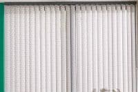 Vertical fabric blinds covering wide window; patterned, closed slats filtering daylight; mounted on top rail with side chain; interior with green left wall and small potted plant.
