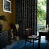 Black patterned floor-to-ceiling curtains cover most of a large sliding door, partially drawn to reveal a sunlit garden; dining area with a wooden chair, set table and a mounted animal skull.