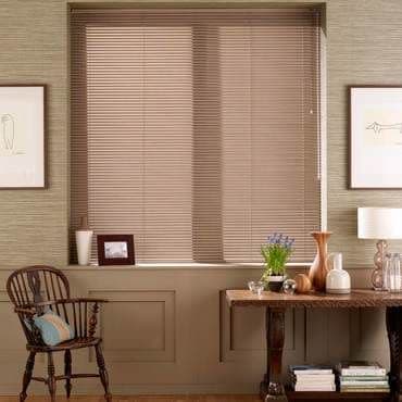 Beige horizontal venetian blinds over a recessed window, fully lowered and closed, gently filtering daylight; surrounding warm sitting area with wooden chair, console table, lamp and small decorative objects.