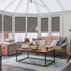 Grey cellular (honeycomb) blinds lowered over tall conservatory windows, softly filtering daylight; cozy sunroom with a beige sofa draped in a chunky pink throw, wooden coffee table and blue rug.