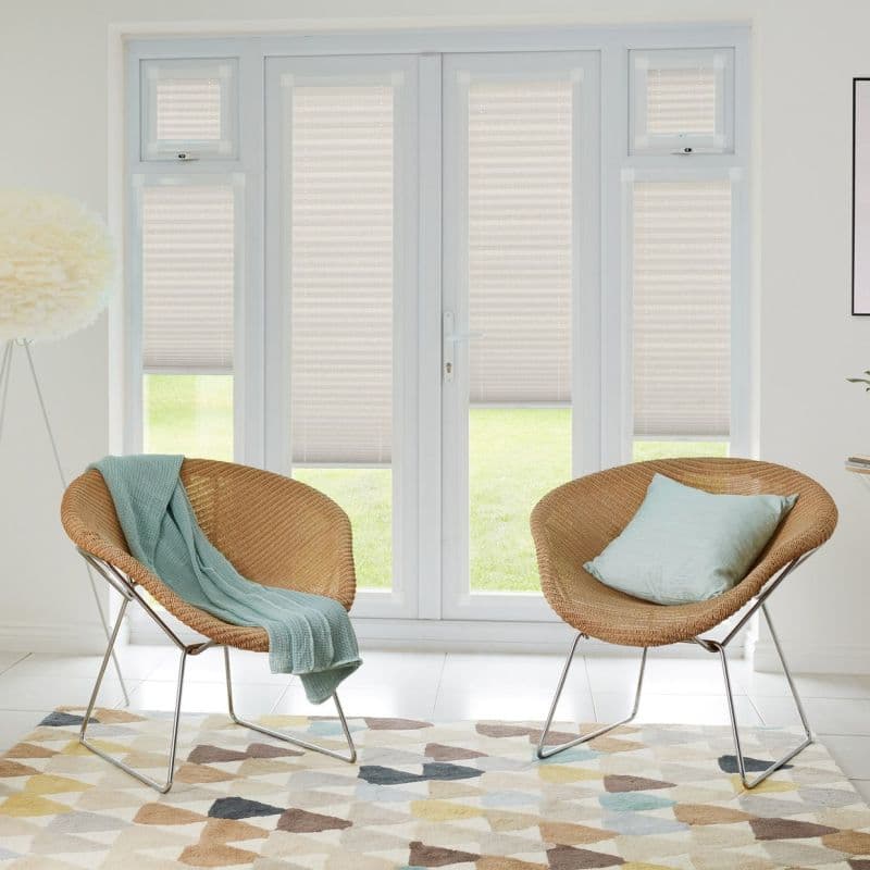Beige pleated light‑filtering shades lowered over double French doors and narrow flanking windows, softly diffusing daylight; bright modern living room with two woven rattan chairs, pastel throw, and patterned rug.