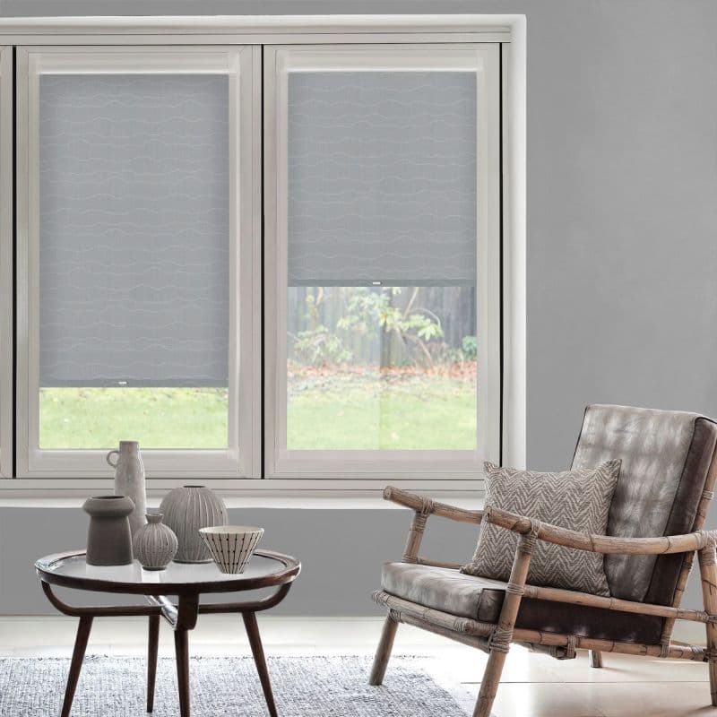 Light-gray patterned roller blinds, lowered partway on twin windows, filtering daylight; neutral-toned living room with rattan-framed leather armchair, patterned cushion and round coffee table with ceramics, grassy yard beyond.