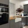White horizontal slatted blinds over two windows, closed and softly filtering light; foreground farmhouse sink and wooden countertop within grey shaker cabinets, stainless ovens and a vase of autumn flowers.
SMEG
23:33