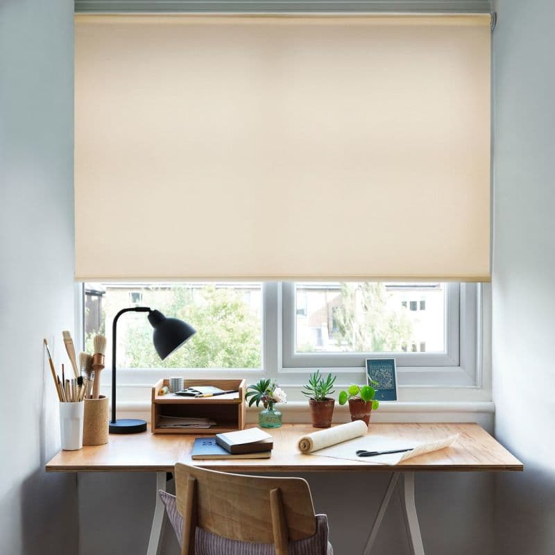 Beige roller blind lowered just above the sill, filtering daylight; beneath, a wooden desk holds a lamp, notebooks, potted succulents and art brushes in a bright home workspace.