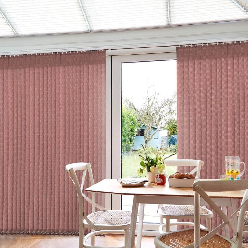 Vertical textured pink blinds, mounted across patio doors, mostly closed with a central gap filtering daylight; dining table and chairs in a bright room, backyard with tree and shed visible.