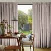 Full-length pale pink textured pinch-pleat curtains, drawn aside to reveal a glass door; soft daylight falls on a wooden dining table set for breakfast with pancakes and wooden chairs.